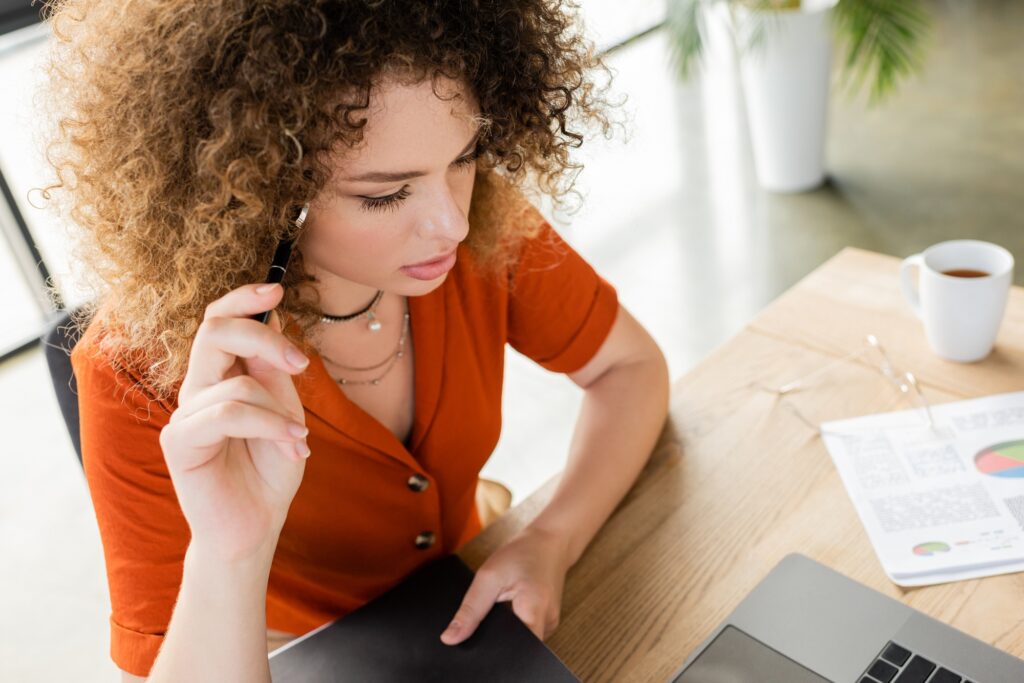 overhead view of pensive businesswoman holding pen and folder near laptop