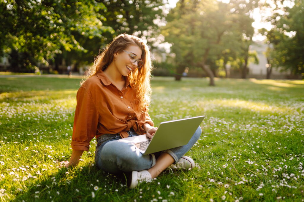 Portrait of a happy woman in sunny park on green lawn with a laptop. Freelance concept, technology.