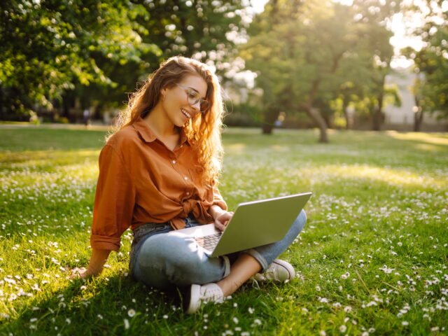 Portrait of a happy woman in sunny park on green lawn with a laptop. Freelance concept, technology.