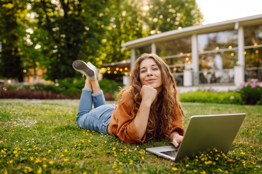 Young curly woman in the park on a green lawn with a laptop in her hands. Online education.