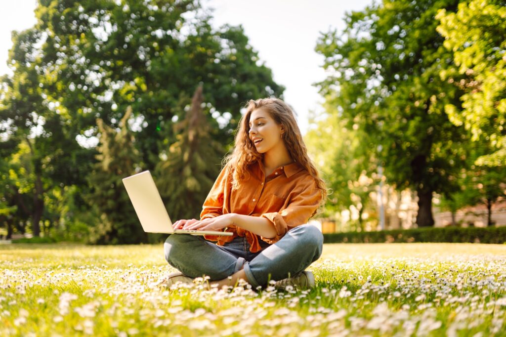 Young curly woman in the park on a green lawn with a laptop in her hands. Online education.